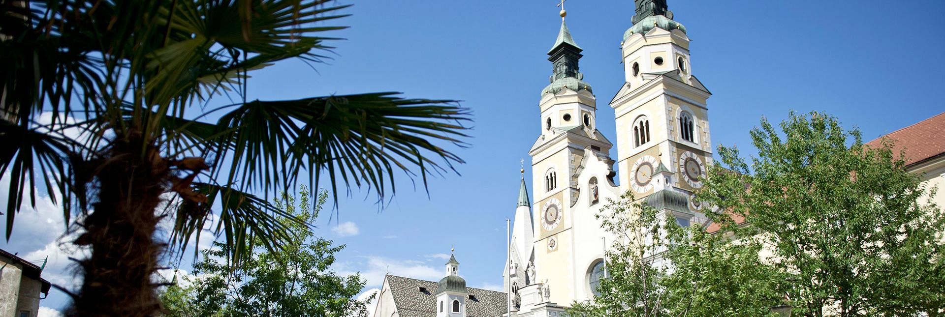 View of the towers of the Bressanone Cathedral, surrounded by palm trees and greenery under a clear blue sky.