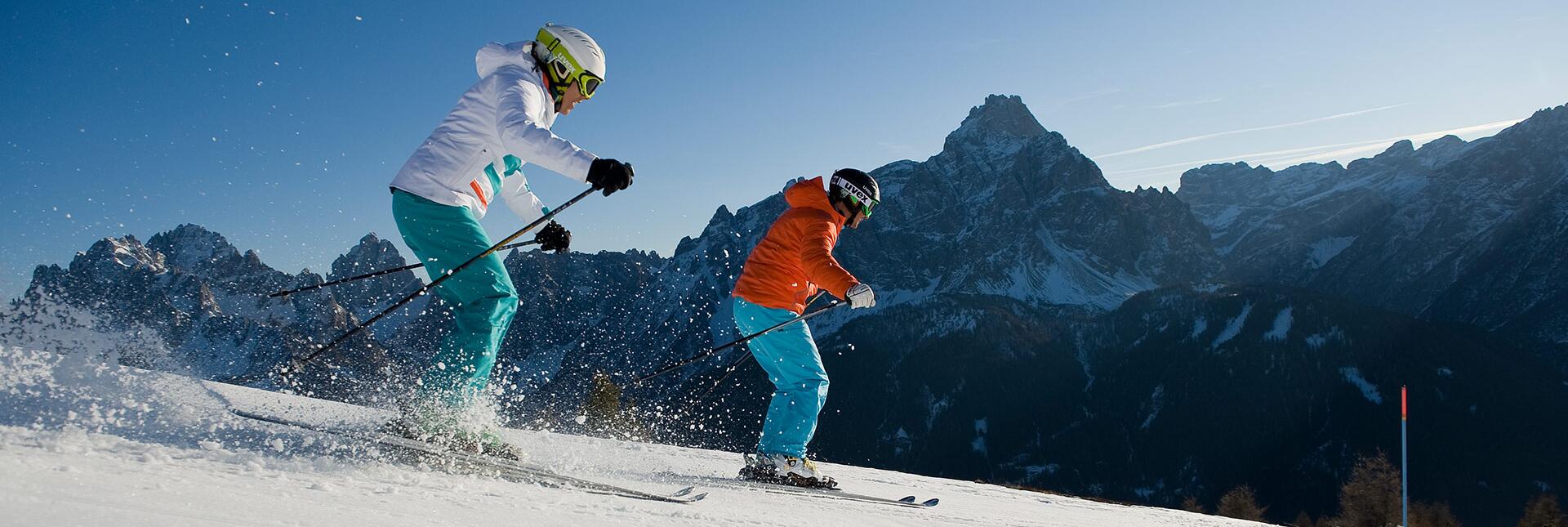 Zwei Skifahrer fahren eine Piste hinab, umgeben von verschneiten Bergen und blauem Himmel.