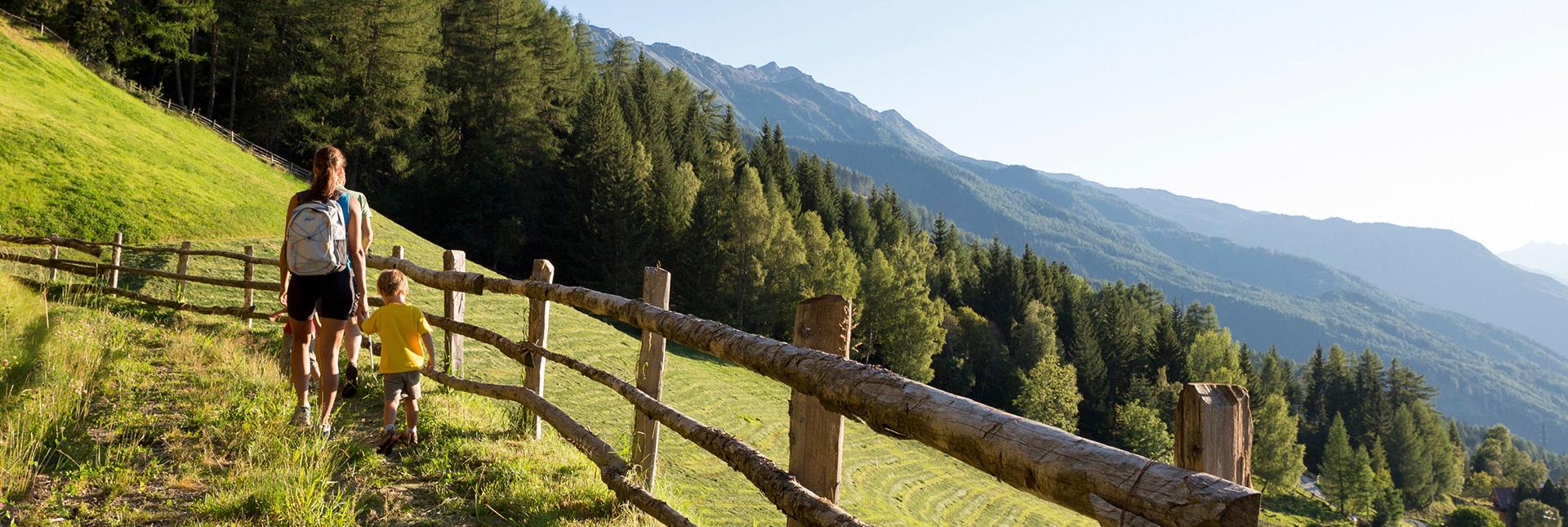 Mother and child hiking on an alpine trail with fence, forest and mountain view at sunset.