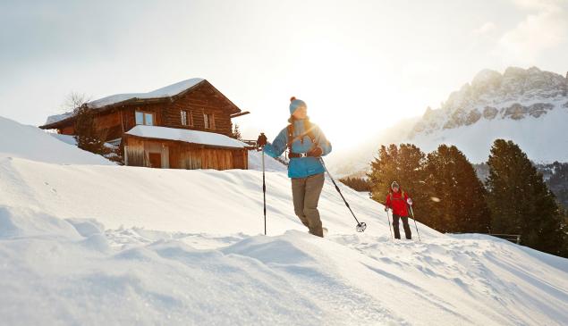 Zwei Personen beim Schneeschuhwandern vor einer Hütte und verschneiten Bergen im Hintergrund.