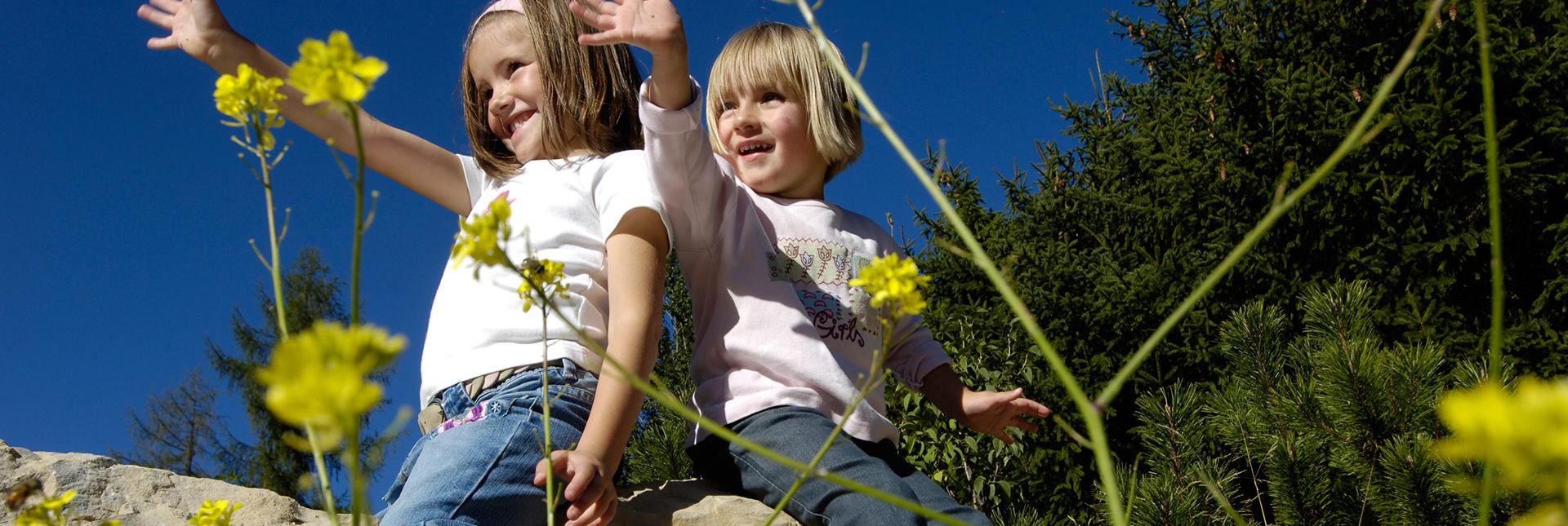 Due bambini sorridenti seduti su una roccia salutano tra fiori gialli e cielo azzurro.